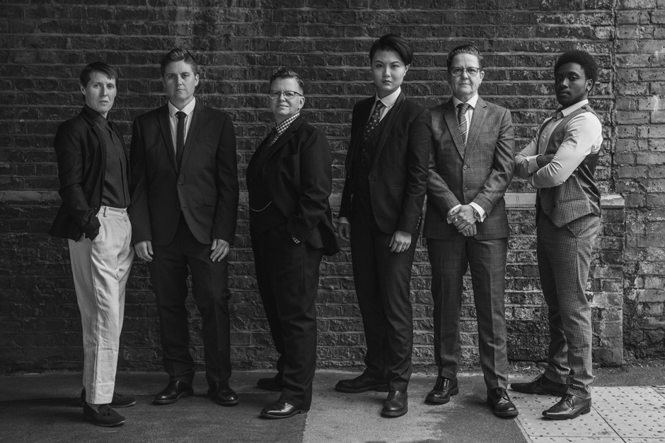 Six people in formal attire stand side by side against a brick wall, posing confidently and looking at the camera in a black-and-white photo.