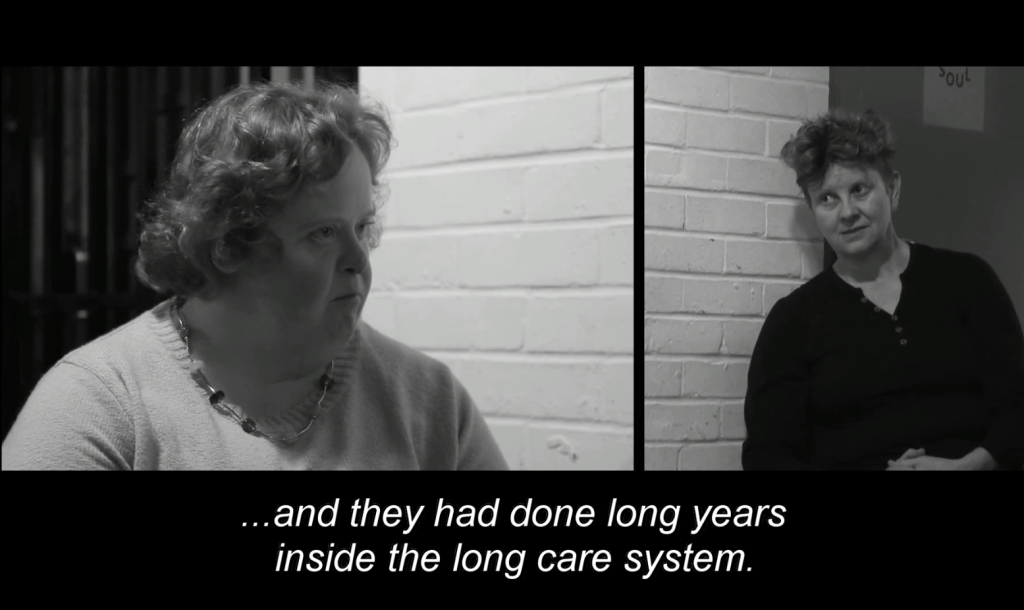 Black and white screenshot from the documentary 'Voices from the Knitting Circle', produced in 2018. Split screen, a blonde woman with short hair on the left (Rachel High) interviewing a woman with short blonde hair on the right (Julie McNamara). Subtitles on screen read :...and they had done long years inside the long care system'.