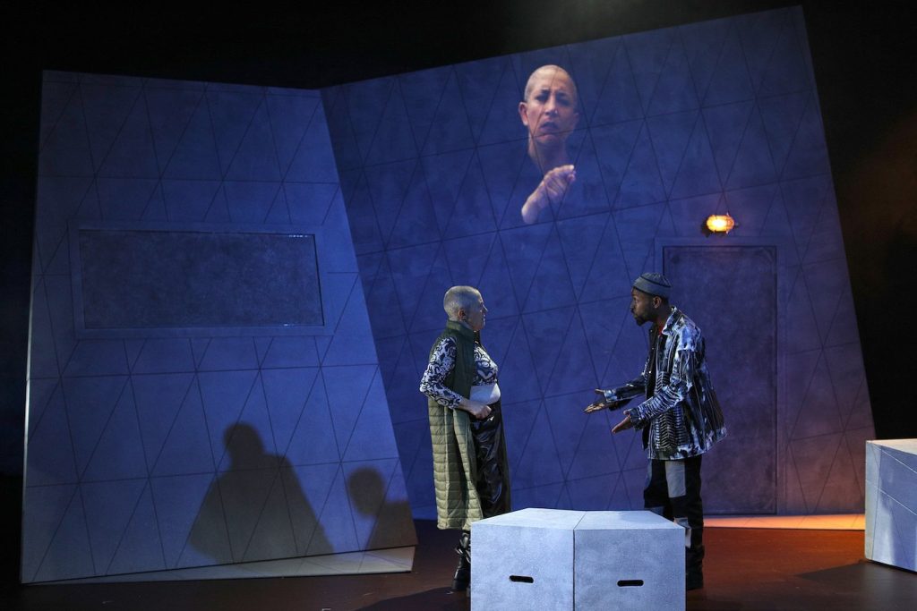 Two people in serious conversation. On the left, a mature white woman with piercing, blue watchful eyes and a shaved head, wearing a green sleeveless coat over a black and white patterned top and black shiny trousers. She is conversing with a young bearded Black man, wearing a black and white patterned hat and jacket. In the background, projected onto a grey wall, a light skinned brown woman with a shaved head interprets their conversation into British Sign Language.