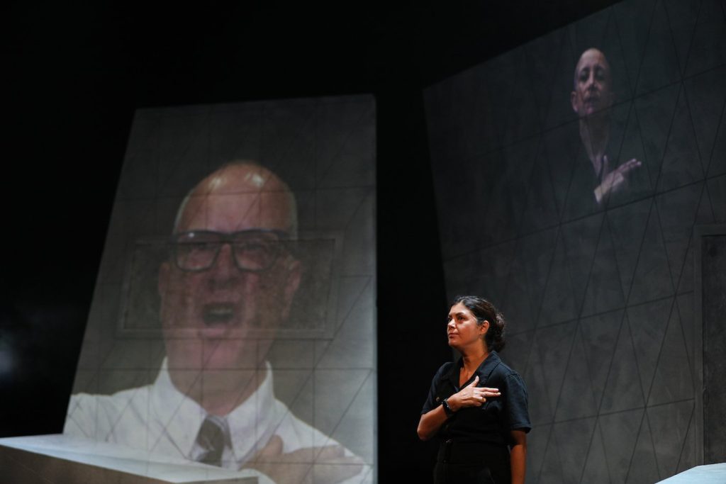 A woman, dressed all in black, stands to attention, her right arm crossed over her chest, with her thumb, index and middle finger resting on her shoulder in a salute. Projected in the background on two large screens, a man with dark framed glasses, white shirt and dark tie, and a woman with a shaved head, dressed in black, both give the same salute.