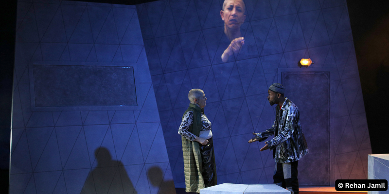 Two people in serious conversation. On the left, a mature white woman with piercing, blue watchful eyes and a shaved head, wearing a green sleeveless coat over a black and white patterned top and black shiny trousers. She is conversing with a young bearded Black man, wearing a black and white patterned hat and jacket. In the background, projected onto a grey wall, a light skinned brown woman with a shaved head interprets their conversation into British Sign Language.