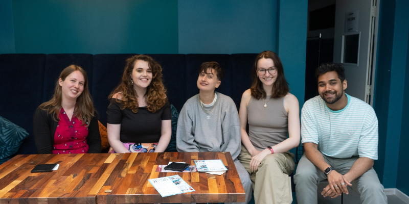 Group photo: five of the Wellspring Writers sitting next to each other, smiling at the camera. From left to right: Emma, Ellie, Jesse, Em and Mohit. Emma: A white woman with multi-coloured eyes and dark blonde straight / slightly wavy hair a few inches beyond her shoulders. Ellie: a woman in her late 20s with brown, curly hair and blue eyes. Jesse: a white androgynous person with short dark hair. Em: they have shoulder length brown hair, a large smile and warm face. Mohit: a short haired South Asian man.