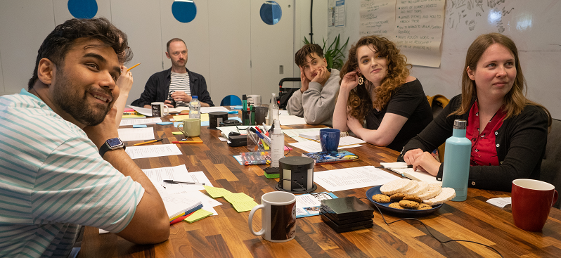 Group photo of the Wellspring training. Six people sit around a table looking to the right of the camera and smiling. Note pads, colourful pens and papers, biscuits and mugs are on the table.