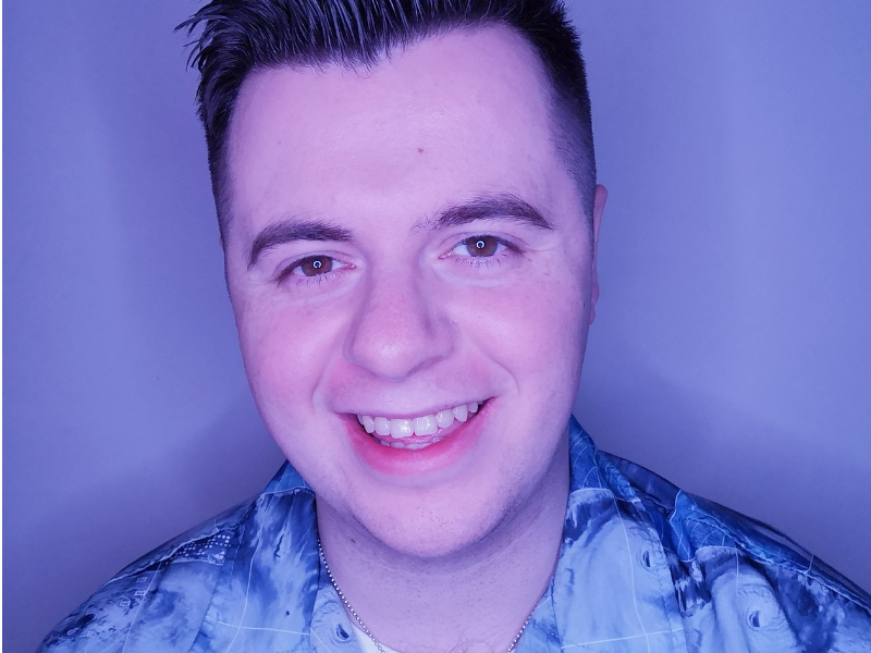 A white man with short dark hair that swoops over to the left. He is wearing a silver-coloured vertical rectangular necklace that rests on a plain white t-shirt, and over the top is a partially buttoned-up shirt that’s a collage of blue and white tones and shows various weather patterns and waves.