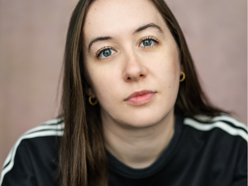 A woman with white skin, blue eyes and long dark brown hair, sat in front of a pink wall. They are wearing a black sweatshirt and gold hoop earrings.