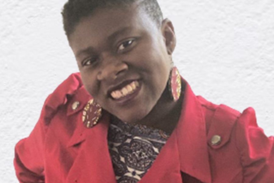 A dark-skinned Black woman smiles and looks at the camera. She is wearing a bright red vintage swallow-tail trench coat with a purple patterned shirt visible underneath, dark red lipstick, and purple patterned earrings. Her hair is shaved on both sides.