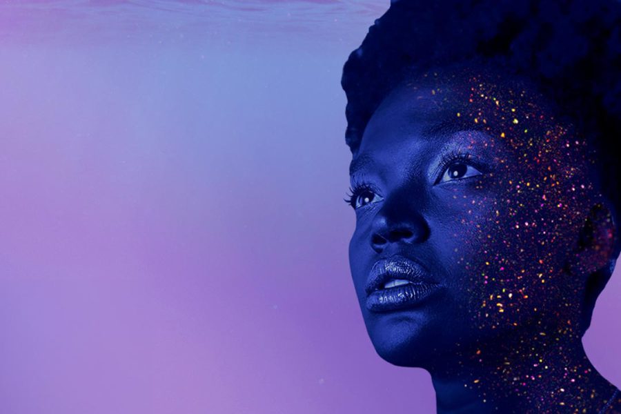 Image of a Black woman’s face, partly covered in sparkling glitter. She intently and blankly gazes outside the frame. The background is filled with gentle water waves in light shades of blue and purple.
