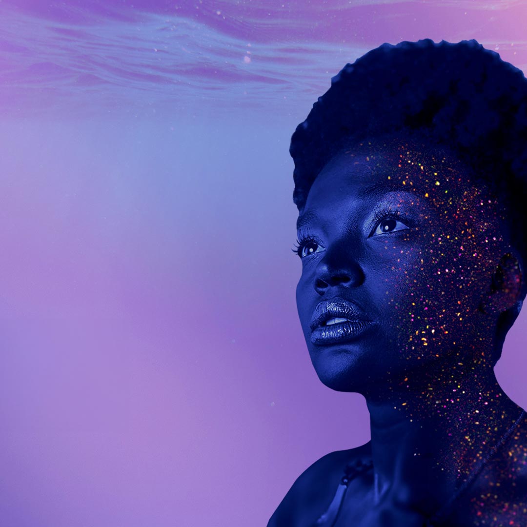 Image of a Black woman’s face, partly covered in sparkling glitter. She intently and blankly gazes outside the frame. The background is filled with gentle water waves in light shades of blue and purple.