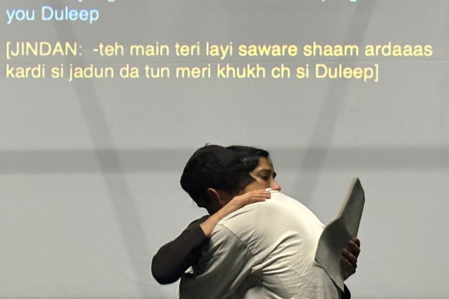 On the left, a South Asian woman with dark hair in bun, holding a script. On the right, a South Asian man wearing a white collarless shirt. They are hugging. On the screen be-hind them, is projected in both English and Punjabi, the words ‘Jindan: -and I have prayed for you every morning and every night from the moment I fell pregnant with you Duleep.’