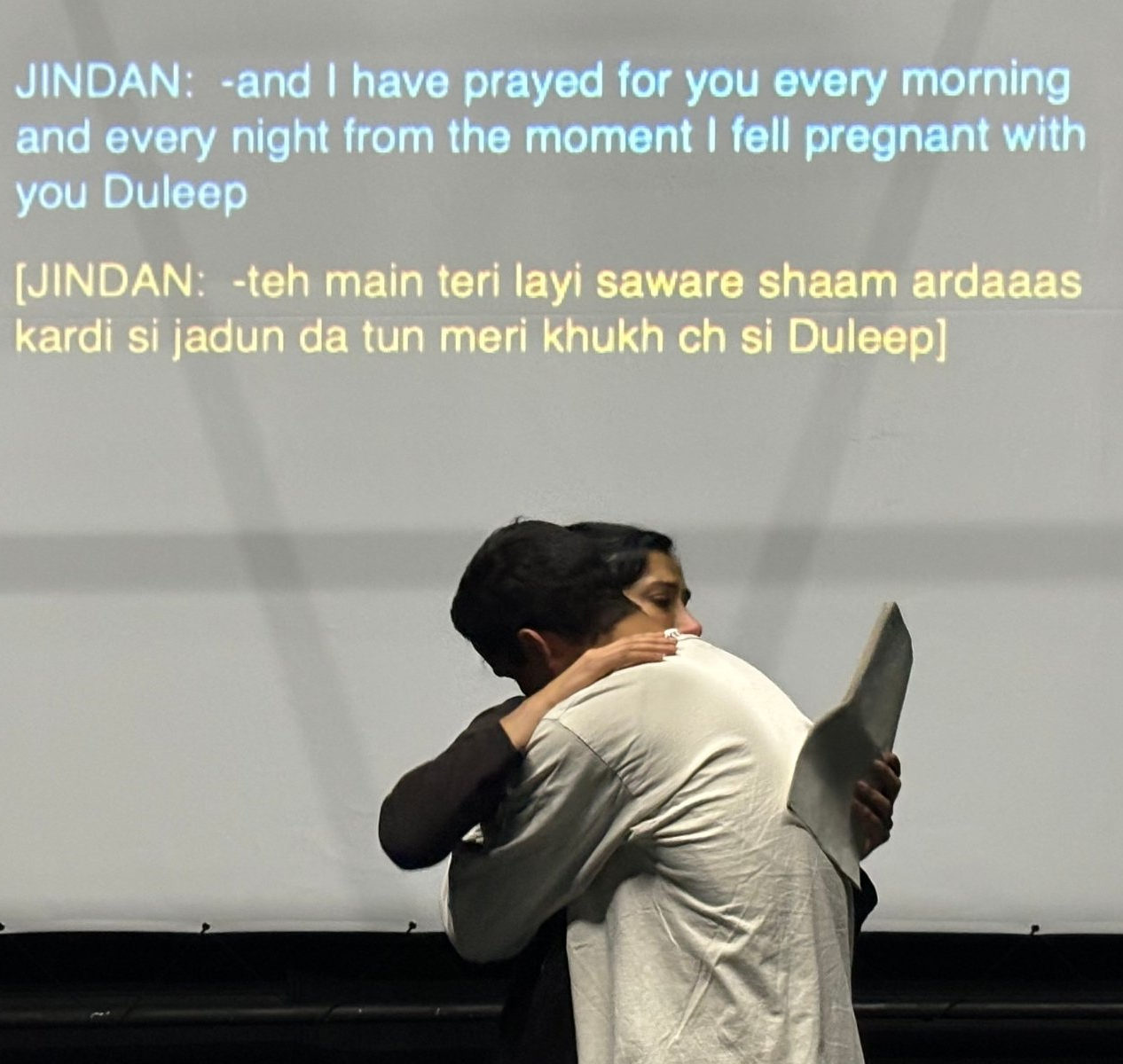 On the left, a South Asian woman with dark hair in bun, holding a script. On the right, a South Asian man wearing a white collarless shirt. They are hugging. On the screen be-hind them, is projected in both English and Punjabi, the words ‘Jindan: -and I have prayed for you every morning and every night from the moment I fell pregnant with you Duleep.’