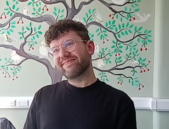 A white man with a short beard and glasses, smiling. He is wearing a black T-shirt. On the wall behind him, a drawing of cherry tree with white birds on its branches.