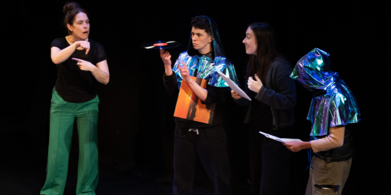 A young person wearing a sparkly metallic hooded cape with a blue cap examining a vinyl record they're holding in one finger. Next to them, a young brunette woman looking at the vinyl record. Near her stands a person wearing a similar sparkly metallic hooded cape, his face doesn't show. On the floor are two shabby old cardboard boxes, two of which contain vinyl records