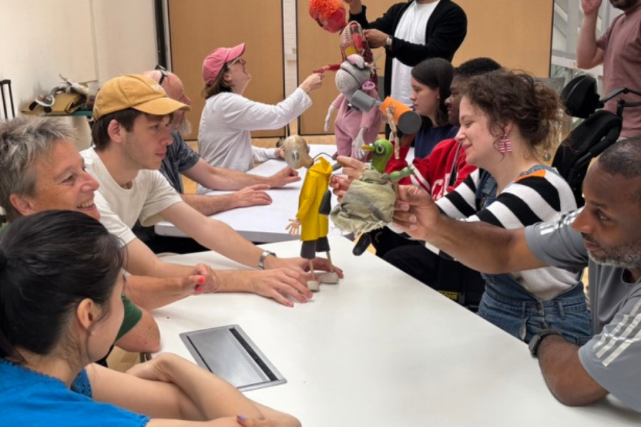 A group of people sit around a white table in a bright room, happily interacting with colourful hand puppets and engaging in conversation.