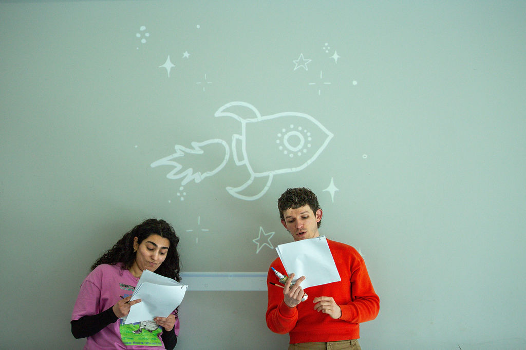 Two people stand under a white rocket drawing on a grey wall, reading from papers. One wears a pink shirt, the other an orange sweatshirt. They both appear focused on the papers they are holding.