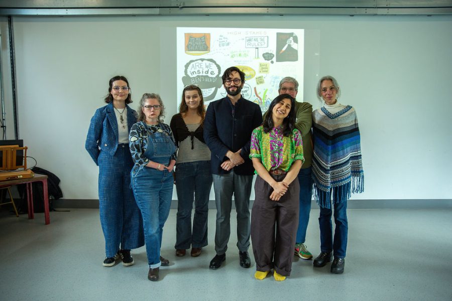 Seven people stand together indoors, smiling at the camera in front of a projection screen displaying colourful, hand-drawn graphics and text.