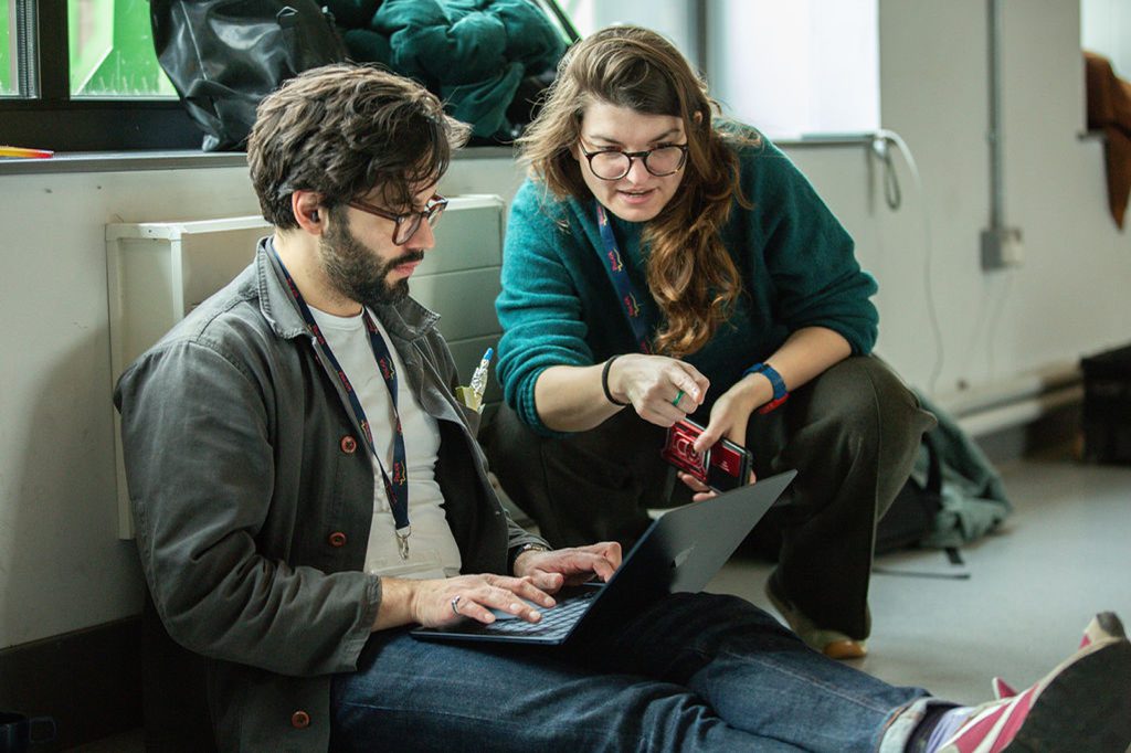 Two people sit on the floor indoors. One types on a laptop while the other crouches beside them, pointing at the screen and holding a wallet. Both wear glasses and appear engaged in discussion.