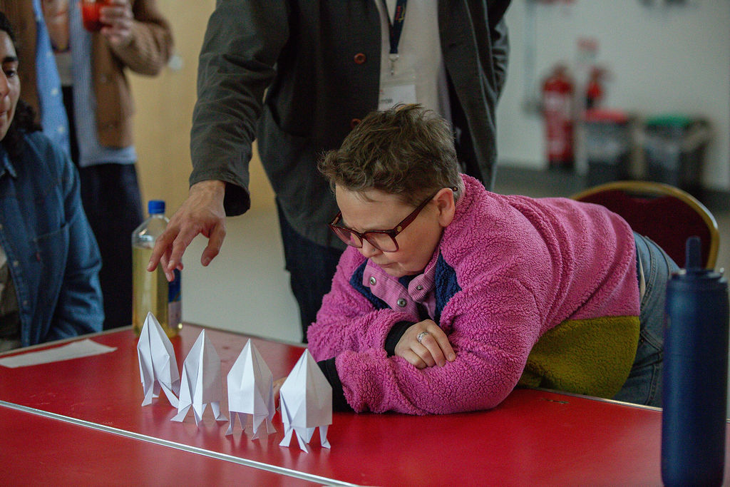 A child in glasses and a pink fleece leans over a red table, looking at four paper rockets. An adult points at the rockets, and other people are partially visible in the background.