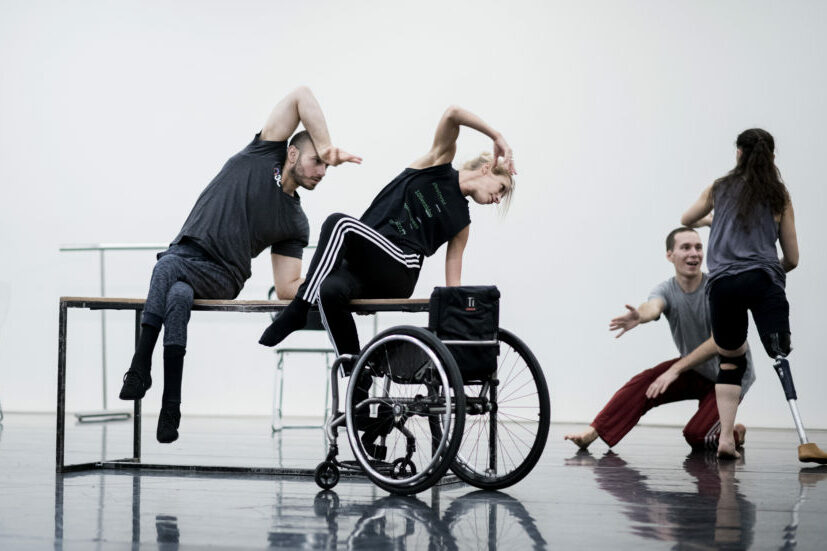 Four dancers, two with visible prosthetic limbs, perform together. One person uses a wheelchair. They are mid-movement on a shiny studio floor, expressing dynamic poses and teamwork.