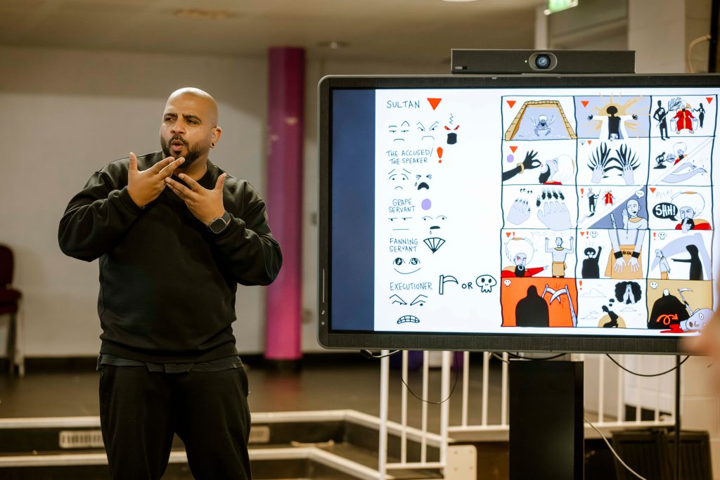 A man in black signs in front of a digital screen displaying colourful comic-style illustrations and text, in a classroom or lecture setting.