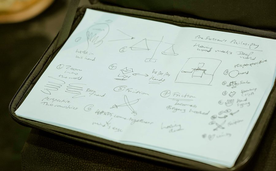 A notebook with hand-drawn sketches and handwritten notes, including arrows, scales, lines, shapes, and brief phrases, lies open on a dark surface.