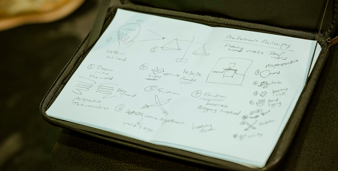 A notebook with hand-drawn sketches and handwritten notes, including arrows, scales, lines, shapes, and brief phrases, lies open on a dark surface.