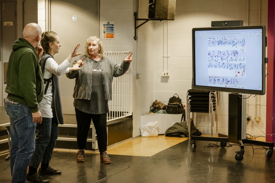 Three people stand in a room. One gestures animatedly whilst another points. A large screen displays illustrated comic panels. Chairs and bags are in the background.