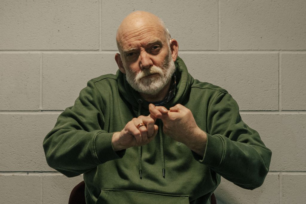 A Deaf bald man with a white beard wearing a green hoodie sits, communicating in sign language.